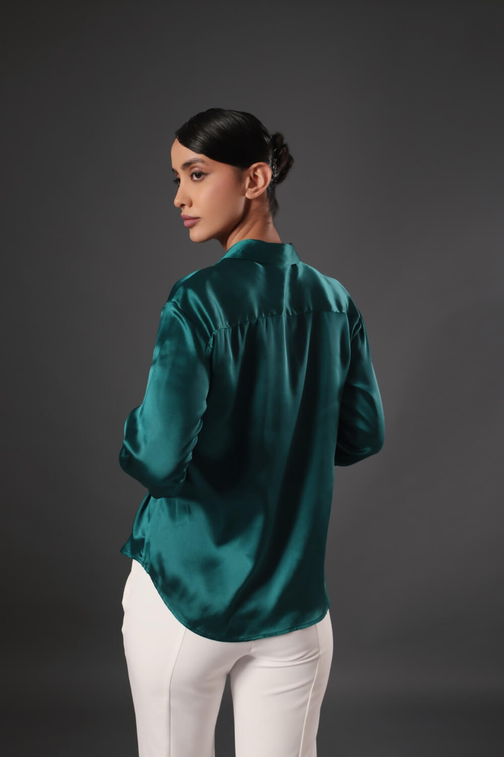 Woman wearing a teal satin blouse and white pants against a dark background