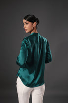 Woman wearing a teal satin blouse and white pants against a dark background