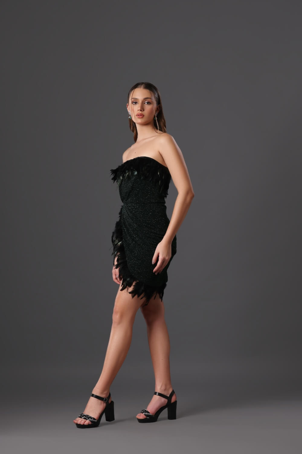 A woman standing in a studio setting, wearing an off-shoulder dark green dress with black feather details, paired with black ankle-strap heels.