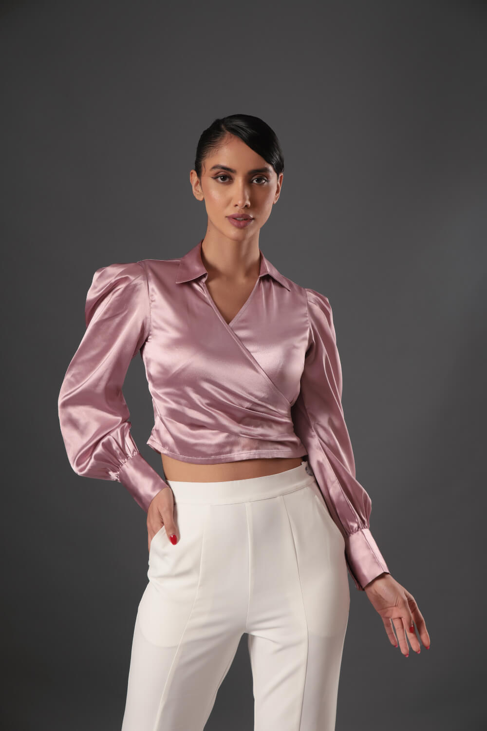 A woman wearing a pink satin top with full sleeves and a wrap design, paired with white trousers.