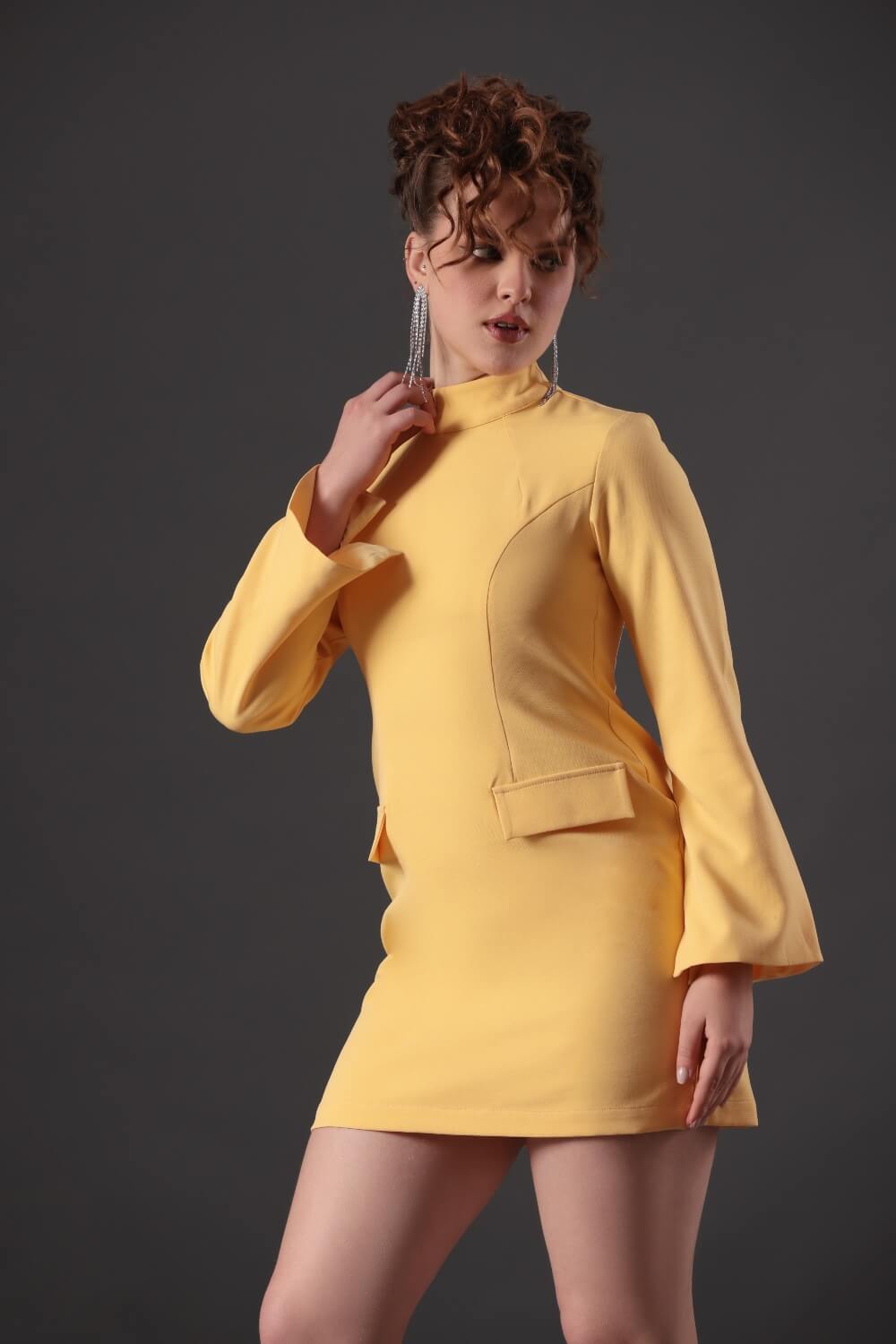 A woman in a yellow high-neck, long-sleeved mini dress with a tailored fit. She has curly hair and is posing with one hand raised to her face.