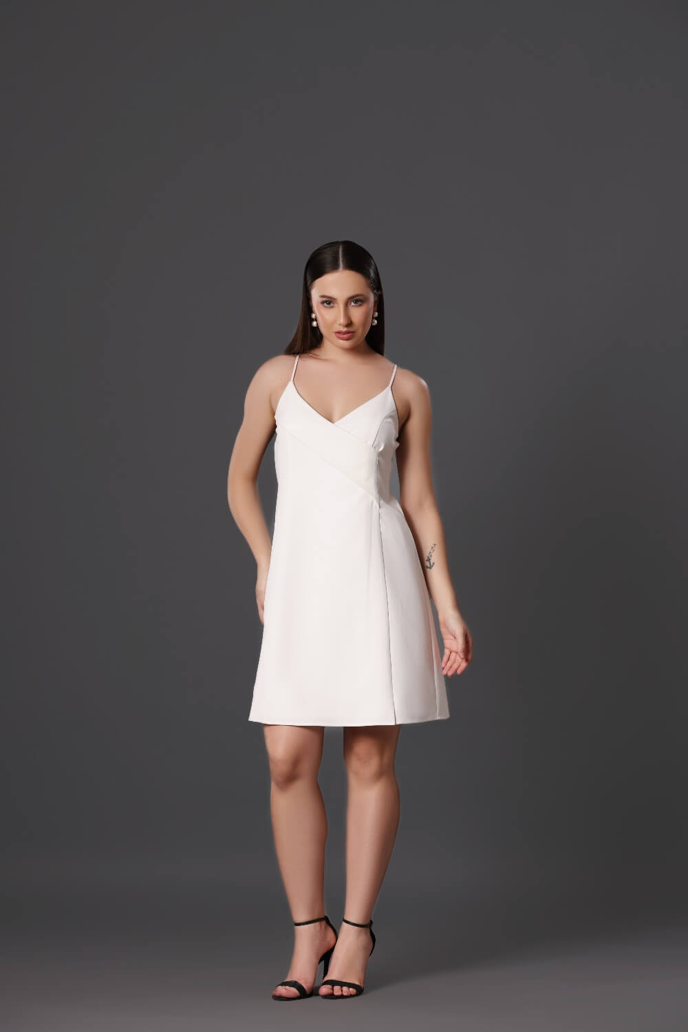 A woman standing in a studio setting, wearing a white sleeveless knee-length dress with a V-neckline and an A-line silhouette. She is also wearing black high-heeled sandals.