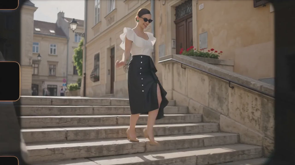 A woman posing in a white ruffled crop top and a black high-low ruffled skirt with silver buttons, standing against a concrete wall.