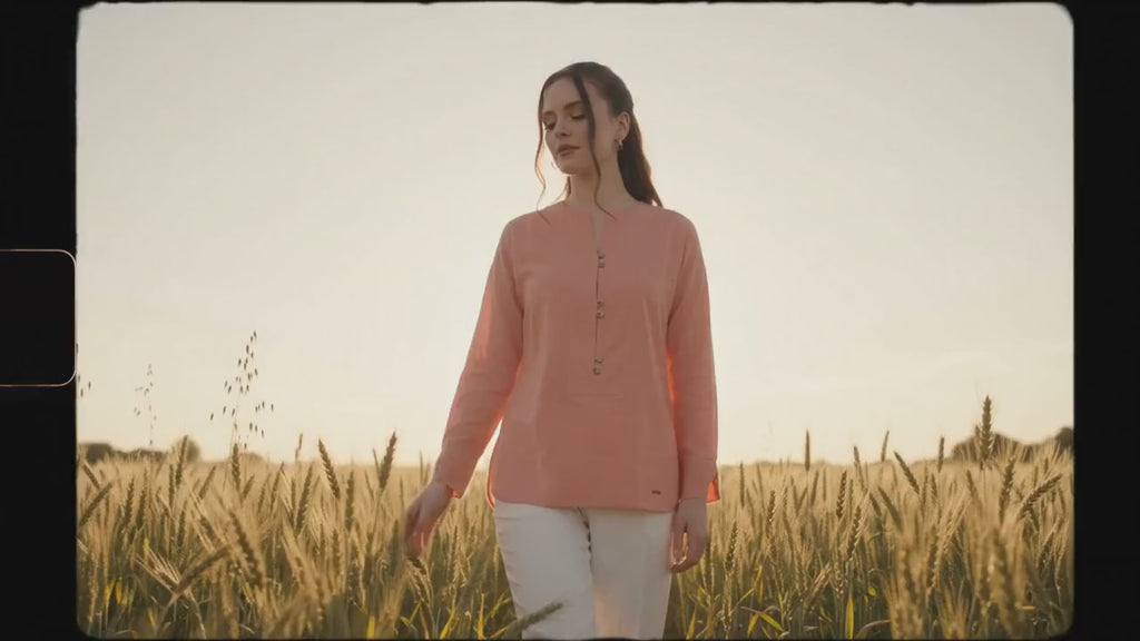 A woman modeling a coral pink long-sleeve button-up top with white trousers and black heels, standing against a dark gray studio background.