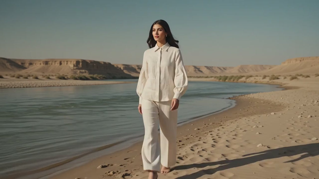 A woman stands in a dry grassy field wearing a white long-sleeve button-up shirt and matching white wide-leg pants.