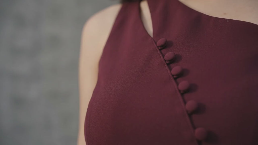 Woman in a burgundy one-shoulder top and wide-leg pants against a gray wall.