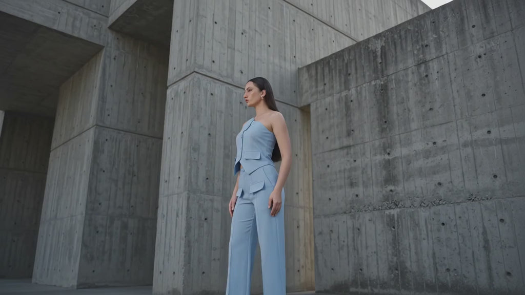 A woman models a light blue one-shoulder top and matching wide-leg trousers set, standing in front of a textured concrete architectural structure with dramatic shadows.