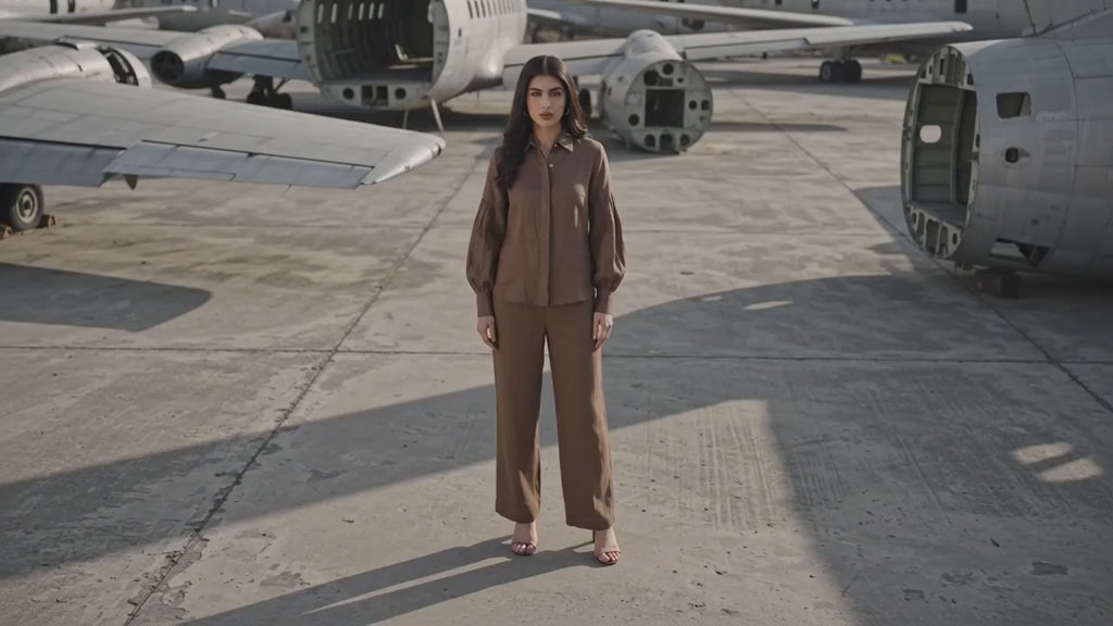 A woman wearing a matching brown long-sleeve button-up shirt and trousers set, leaning against a textured concrete pillar in an industrial setting.