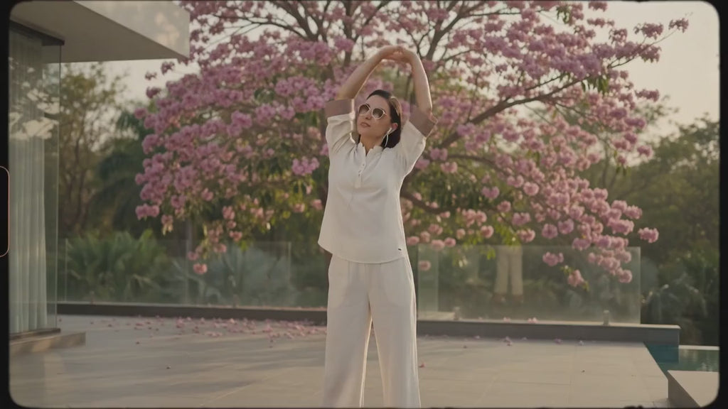 A woman modeling a two-piece cream-colored outfit consisting of a short-sleeve button-up kurta with brown contrast cuffs and matching wide-leg trousers with brown contrast hems, set against a solid dark gray studio background.