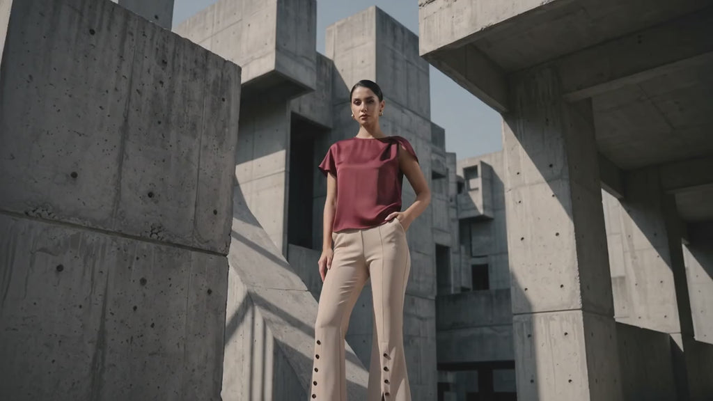 A woman modeling a short-sleeve burgundy top and beige flared trousers with side buttons, standing in front of a modern concrete building.